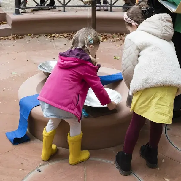 2 Kinder mit dem Rücken zur Kamera und ein Pfarrer beim Auftsellen der Taufschalen. Im Hintergrund weitere Kinder und Erwachsene, die an einem Geländer stehen.