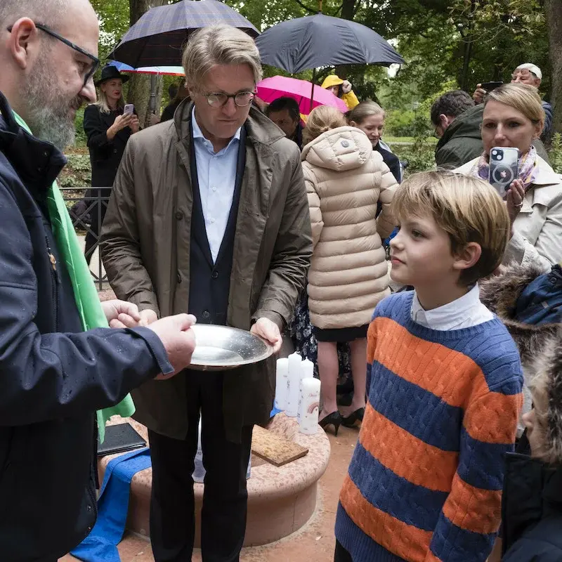 Eine Familie beim Tauffest. Im Vordergrund der Pfarrer, der Täufling und der Taufpate, der die Taufschale in seinen Händen hält. Dahinter die fotografierende Mutter des Täuflings. Außerdem weitere Besucher*innen mit Schirmen.